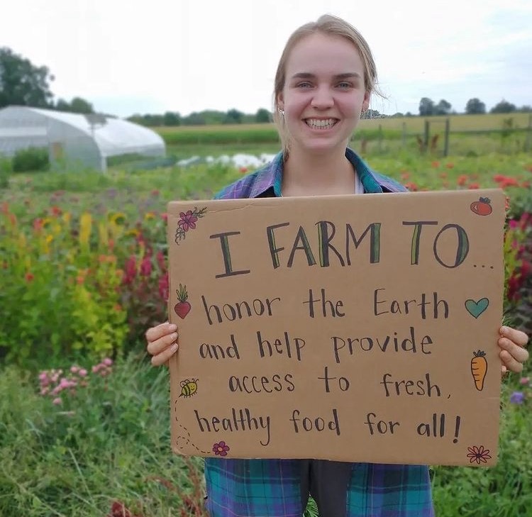 Maria Bossert holding up a sign that reads I farm to... honor the Earth and help provide access to fresh, healthy food for all!
