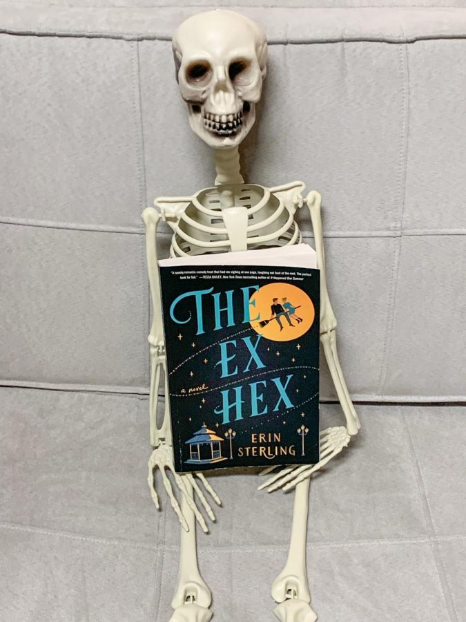 A skeleton holding a copy of The Ex Hex book.